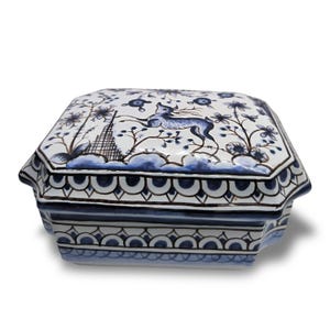Könnte beinhalten: Achteckige Schmuckschatulle aus Keramik mit Deckel. Die Box ist weiß mit blauen Blumen- und Hirschmotiven. Der Rand und die Basis weisen ein sich wiederholendes blaues und braunes Muster auf. Die Box ist wahrscheinlich handbemalt.
