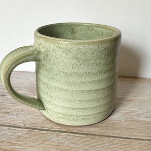 May include: A handmade ceramic mug in a soft green hue. The mug features a rounded body with a sturdy handle and a textured, speckled surface. The design includes horizontal bands, adding visual interest to the piece. A simple, functional piece for enjoying beverages.
