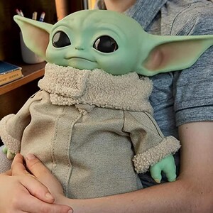 May include: A green creature figurine with large black eyes and pointed ears, wearing a tan jacket with a textured cream-colored sherpa collar and matching cuffs on the sleeves. The figure has a smooth skin texture and a neutral facial expression.