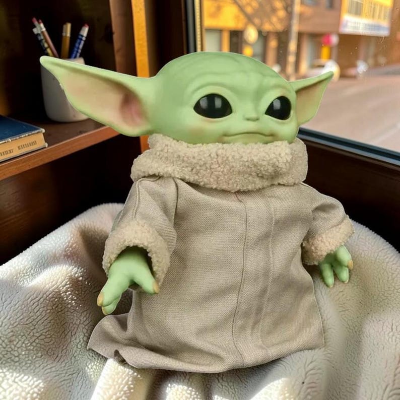 May include: A green creature figurine with large ears and black eyes, wearing a beige robe with a textured cream-colored sherpa collar and sleeve cuffs. The figure has a smooth skin texture and is positioned in a seated posture with arms extended.