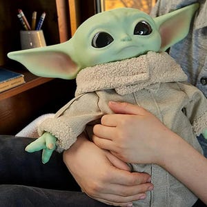 May include: A green creature figure with large black eyes and pointed ears, wearing a beige jacket with a textured cream-colored sherpa collar and cuffs. The figure has a smooth, matte skin finish and is depicted in a seated position.