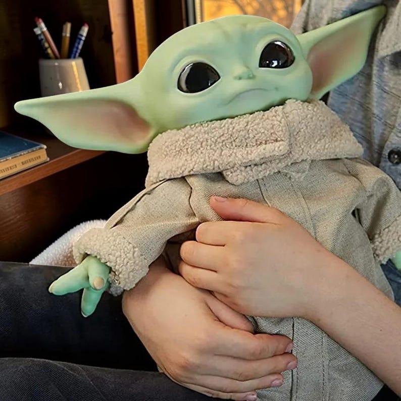 May include: A green creature figure with large black eyes and pointed ears, wearing a beige jacket with a textured cream-colored sherpa collar and cuffs. The figure has a smooth, matte skin finish and is depicted in a seated position.