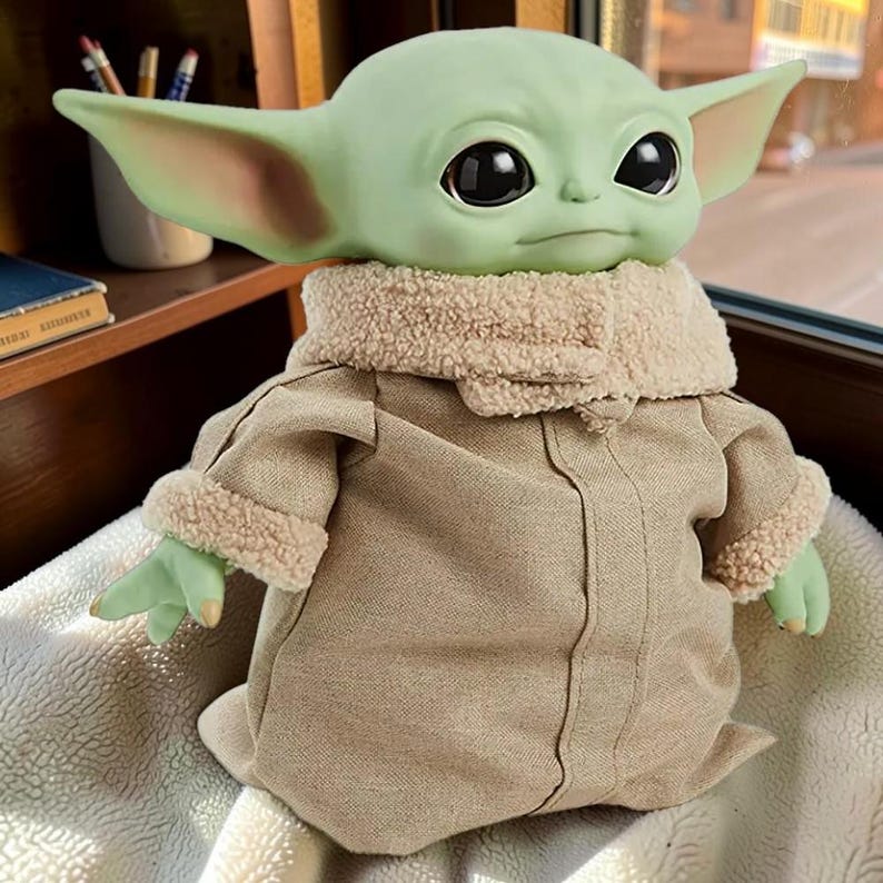 May include: A green creature figurine with large black eyes and pointed ears, wearing a tan robe with a textured beige faux-shearling collar and matching sleeve cuffs. The figure has a smooth skin texture and is positioned in a seated posture.