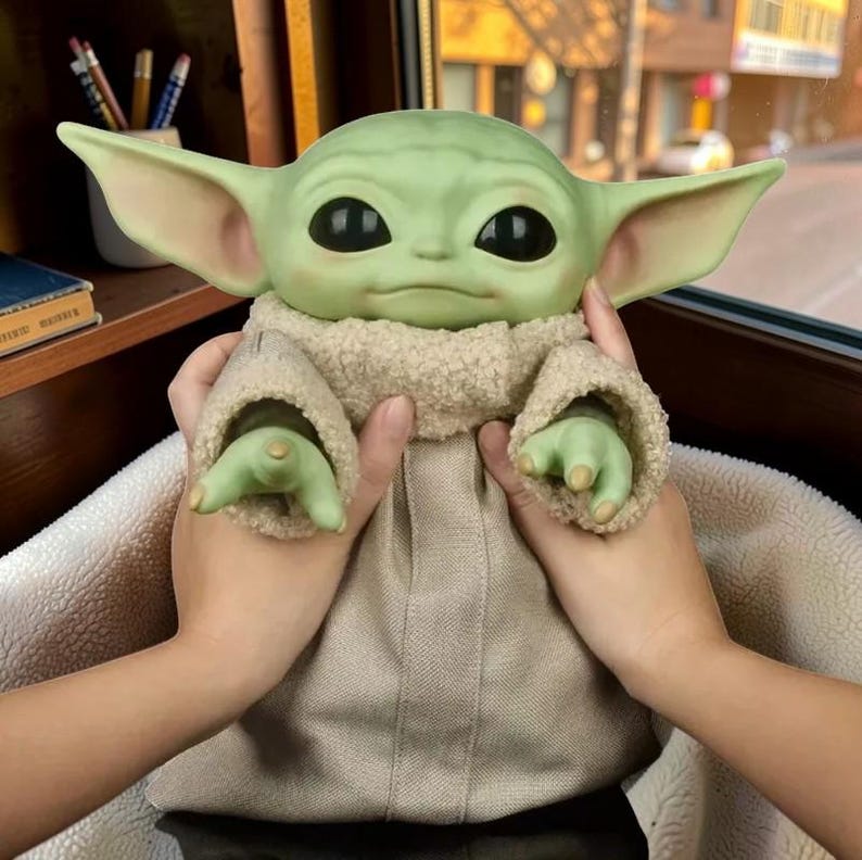 May include: A green creature figurine with large black eyes, pointed ears, and a wide head. The figure wears a beige textured robe with a fuzzy, cream-colored collar that wraps around the neck and extends down the front of the garment.