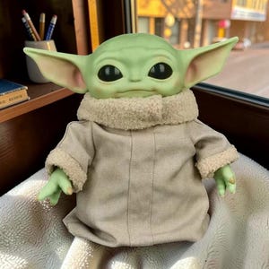 May include: A green creature figurine with large pointed ears and black eyes, wearing a beige robe with a textured cream-colored faux-fur collar and sleeve cuffs. The figure has a smooth, rounded head and small hands extending from the sleeves.