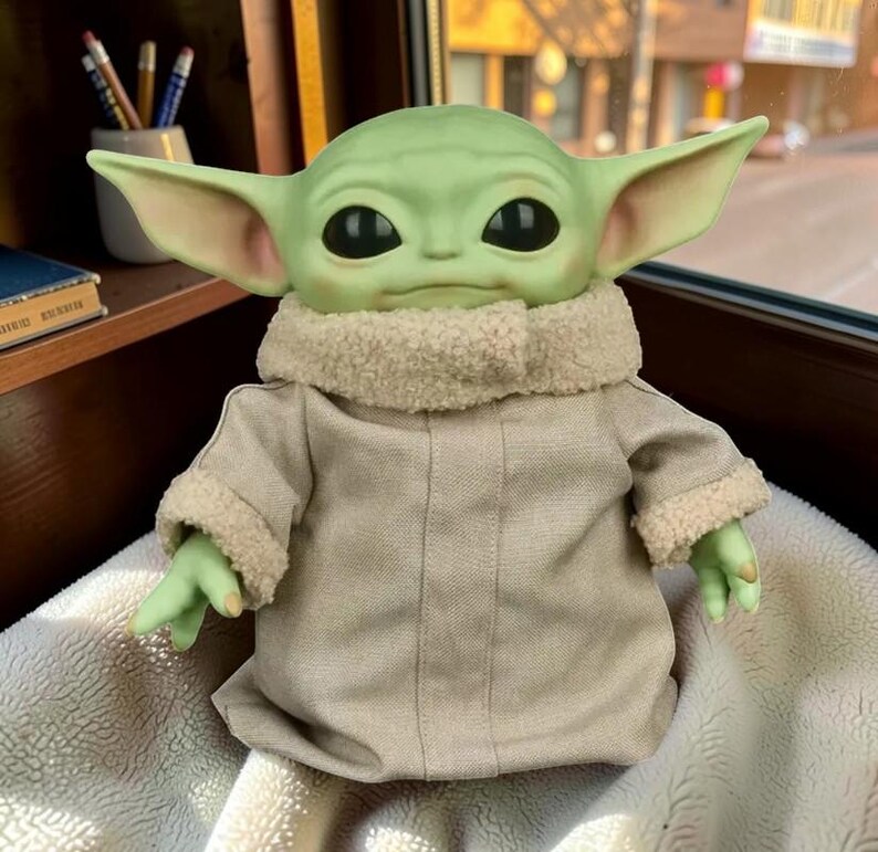 May include: A green creature figurine with large pointed ears and black eyes, wearing a beige robe with a textured cream-colored faux-fur collar and sleeve cuffs. The figure has a smooth, rounded head and small hands extending from the sleeves.