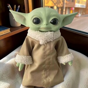 May include: A green creature figurine with large black eyes and pointed ears, wearing a tan robe featuring a white fuzzy collar and white fuzzy cuffs on the sleeves. The figure has a smooth, light green skin texture and a neutral facial expression.