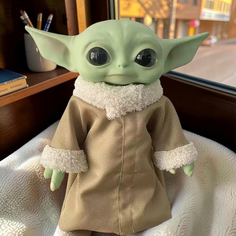May include: A green creature figurine with large black eyes and pointed ears, wearing a tan robe featuring a white fuzzy collar and white fuzzy cuffs on the sleeves. The figure has a smooth, light green skin texture and a neutral facial expression.