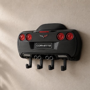 May include: A black wall-mounted key holder shaped like the rear end of a Corvette sports car. It features four red circular taillights, a central Corvette emblem, a license plate area labeled CORVETTE, and four black hooks extending downward.