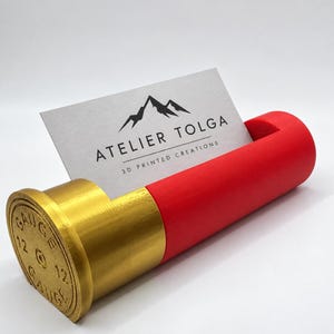Op de afbeelding: Een 3D-geprinte visitekaarthouder in de vorm van een 12 gauge jachtpatroon met een metallic gouden basis en een matrode behuizing. Een wit visitekaartje zit in de gleuf, met een berglogo en de tekst ATELIER TOLGA 3D PRINTED CREATIONS.