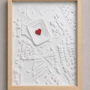 Op de afbeelding: Een ingelijste 3D-reliëfkaart in wit met een getextureerd stadslandschap inclusief een stadion. Een klein, rood hartje is in het midden van het stadion geplaatst. Het kunstwerk zit in een lichtgekleurde houten lijst met een strak, rechthoekig ontwerp.