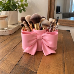 May include: A light pink cylindrical makeup brush holder featuring a prominent decorative bow on the front. The holder is filled with various makeup brushes of different sizes and bristle types, including fluffy powder brushes and thin eyeliner brushes.