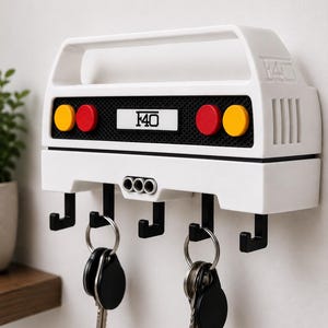 Peut inclure: Un porte-clés mural blanc en forme de l'arrière d'une voiture de sport F40. Il présente une grille noire, deux feux rouges et deux jaunes, ainsi que le texte F40 sur la plaque centrale. Cinq crochets noirs soutiennent deux trousseaux de clés.