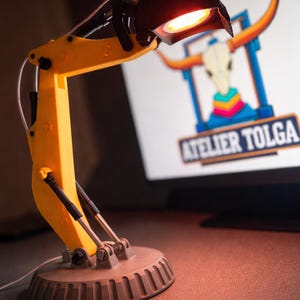 Op de afbeelding: Een gele bureaulamp met een zwarte kap, ontworpen om op een graafmachinearm te lijken. De lamp heeft een grijze voet en is verlicht. De achtergrond toont een logo met een schedel van een stier en de tekst "ATELIER TOLGA".