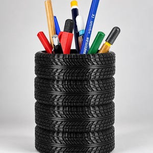 Op de afbeelding: Een zwarte pennenhouder in de vorm van een stapel van vier banden, gevuld met diverse schrijfinstrumenten waaronder een blauw Staedtler ergosoft potlood, een houten potlood, een rode stift, een groene stift en andere diverse pennen en stiften.