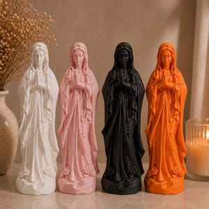 Op de afbeelding: Vier beelden van de Maagd Maria in wit, roze, zwart en oranje. Elk beeld heeft een rozenkrans en staat op een voet met rozen. De beelden zijn in een rij op een reflecterend oppervlak geplaatst.