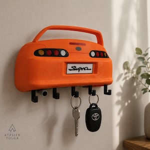 Könnte beinhalten: Ein orangefarbener Wand-Schlüsselhalter in Form des Hecks eines Toyota Supra, mit Spoiler, runden Rückleuchten und einem Nummernschild mit der Aufschrift Supra. Er verfügt über fünf schwarze Haken, an denen ein Schlüssel und ein Toyota-Autoschlüssel hängen.