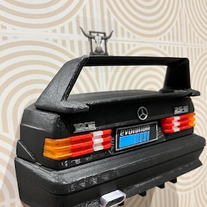 Può includere: Modello nero stampato in 3D di un'auto Mercedes-Benz 190E Evolution. Il modello presenta uno spoiler posteriore, fanali posteriori e la scritta "evolution". Una piccola statuetta a forma di teschio di toro è posta sopra l'auto.