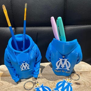 Könnte beinhalten: Zwei blaue Stifthalter in Form von Kapuzenpullovern, jeweils mit weißem Logo. Einer enthält Pinsel, der andere Stifte. Darunter zwei passende Schlüsselanhänger. Der Text "DROIT AU BUT" befindet sich auf der Rückseite eines Halters.