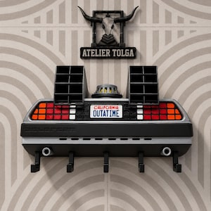 Puede incluir: Un llavero de pared diseñado como la parte trasera de un coche DeLorean, con luces traseras rojas y blancas, una matrícula California OUTATIME, dos rejillas de ventilación negras y cinco ganchos negros. Arriba hay un letrero negro ATELIER TOLGA.