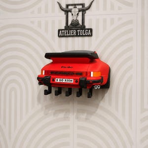 May include: A red Porsche Turbo rear-end wall-mounted coat rack featuring a black spoiler, taillights, and a number plate reading S GO 930H. Below the bumper are five black hooks. Above the car is a black sign with a bull skull and ATELIER TOLGA.
