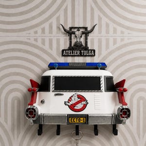 Op de afbeelding: Een wandgemonteerde sleutelhouder in de vorm van de achterkant van het Ecto-1 voertuig, met een witte carrosserie, rode staartvinnen, een blauwe lichtbalk en het Ghostbusters-logo. Onder de bumper zitten vijf zwarte haken. Erboven een bord ATELIER TOLGA.