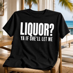 Op de afbeelding: Zwart T-shirt met de witte tekst "LIQUOR? YA IF SHE'LL LET ME". Het shirt heeft een ronde hals en wordt gepresenteerd op een houten stoel. Een glas water met een schijfje limoen staat op een tafel op de achtergrond.