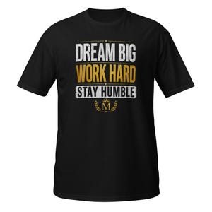 May include: Black t-shirt with the text "DREAM BIG WORK HARD STAY HUMBLE" in white and gold lettering. The design includes a crown and laurel wreath graphic. The shirt is a classic crew neck style.