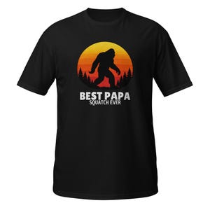 Può includere: T-shirt nera con una silhouette di Bigfoot davanti a un tramonto. Il testo "BEST PAPA SQUATCH EVER" è stampato in bianco. Il design presenta un Bigfoot che cammina davanti a un tramonto. La maglietta è in materiale morbido.