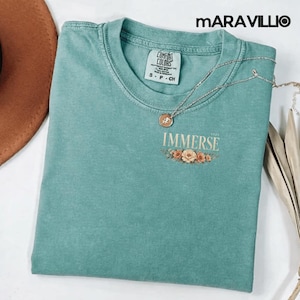 Könnte beinhalten: Ein teal Comfort Colors T-Shirt mit dem Wort "IMMERSE" in Gold gedruckt, mit einem floralen Design darunter. Eine silberne Halskette mit Anhänger ist über dem Shirt drapiert. Das Shirt ist Größe S-P-CH.