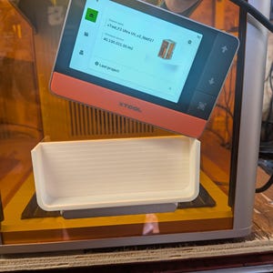 Könnte beinhalten: Ein 3D-Drucker mit einem orangefarbenen und schwarzen Touchscreen, der Geräteinformationen anzeigt. Unter dem Bildschirm befindet sich eine weiße Ablage. Der Drucker ist silberfarben und hat eine transparente Oberseite. Der Text "XTool F2 Ultra UV_v2_5bbf27" wird angezeigt.