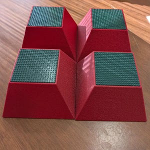 Puede incluir: Cuatro soportes rojos con forma de pirámide dispuestos en formación cuadrada. Cada soporte presenta una superficie superior plana y cuadrada con una almohadilla de agarre antideslizante texturizada de color verde oscuro. Los lados son inclinados.