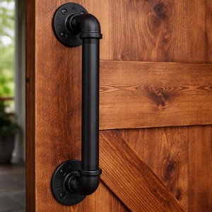 May include: Black industrial door pull handle fixed to a wooden door. The handle is constructed from black pipes and fittings, with circular mounting plates. The door features a warm, brown wood grain finish.