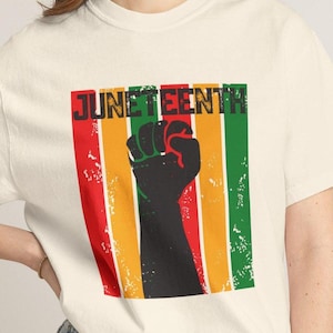 May include: Cream-colored t-shirt featuring a graphic design with the word "JUNETEENTH" in black letters above a black fist. The design includes vertical stripes in red, yellow, and green.