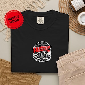 May include: Black t-shirt with a graphic that says "Hustle" in red and white, with a cityscape design. The shirt has a crew neck and short sleeves. A red sticker with "Hustle Mood" is in the upper left corner.