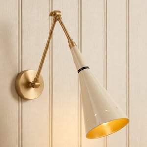 Può includere: Una lampada da parete con braccio in ottone dotato di cerniera regolabile e paralume conico color bianco sporco. Il paralume presenta una sottile fascia nera vicino alla sommità e piccole perforazioni circolari, con una finitura interna dorata.