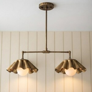 May include: A dual-light pendant fixture featuring a brass-toned metal finish with a central vertical rod and a horizontal bar. Two ruffled, scalloped-edge metal shades hang from the ends, each housing a round, frosted white glass light bulb.