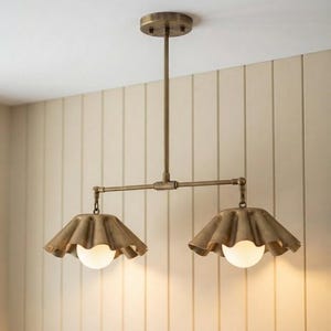 May include: A dual-light ceiling fixture featuring a brass finish with a central rod and two horizontal arms. Each arm supports a wide, ruffled, bell-shaped metal shade in a matching brass tone, housing a single round, frosted white glass bulb.