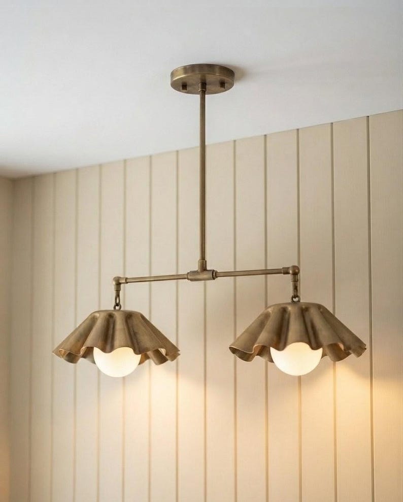 May include: A dual-light ceiling fixture featuring a brass finish with a central rod and two horizontal arms. Each arm supports a wide, ruffled, bell-shaped metal shade in a matching brass tone, housing a single round, frosted white glass bulb.