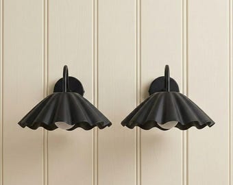 May include: A pair of wall-mounted light fixtures featuring matte black metal shades with a scalloped, fluted edge design. Each fixture includes a curved arm extending from a circular wall plate and a single white bulb visible underneath.