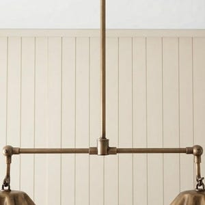 May include: A brass-toned metal pendant light fixture featuring a central vertical rod connected to a horizontal bar. Two articulated arms extend from the bar, each supporting a fluted, bell-shaped shade with a metallic finish and visible joints.