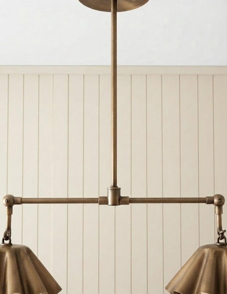 May include: A brass-toned metal pendant light fixture featuring a central vertical rod connected to a horizontal bar. Two articulated arms extend from the bar, each supporting a fluted, bell-shaped shade with a metallic finish and visible joints.