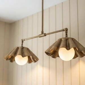 May include: A dual-pendant light fixture featuring a horizontal brass rod suspended from a central stem. Two brass shades with a scalloped, ruffled edge design hang from the rod, each housing a round, glowing white glass globe light bulb.