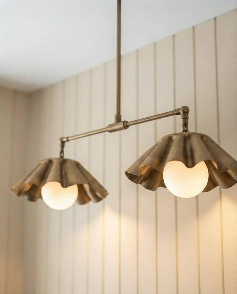 May include: A dual-pendant light fixture featuring a horizontal brass rod suspended from a central stem. Two brass shades with a scalloped, ruffled edge design hang from the rod, each housing a round, glowing white glass globe light bulb.