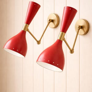 Puede incluir: Dos apliques de pared rojos con detalles en latón. Cada aplique tiene una pantalla en forma de cono con pequeños agujeros y un brazo de latón que se conecta a un soporte de pared redondo de latón. Están montados en una pared clara con paneles verticales.