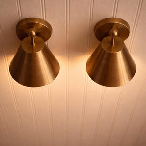 May include: Two brass-colored cone-shaped wall sconces mounted on a white paneled ceiling. Each sconce has a circular base and a small, spherical detail at the top. The sconces are illuminated, casting a warm glow.