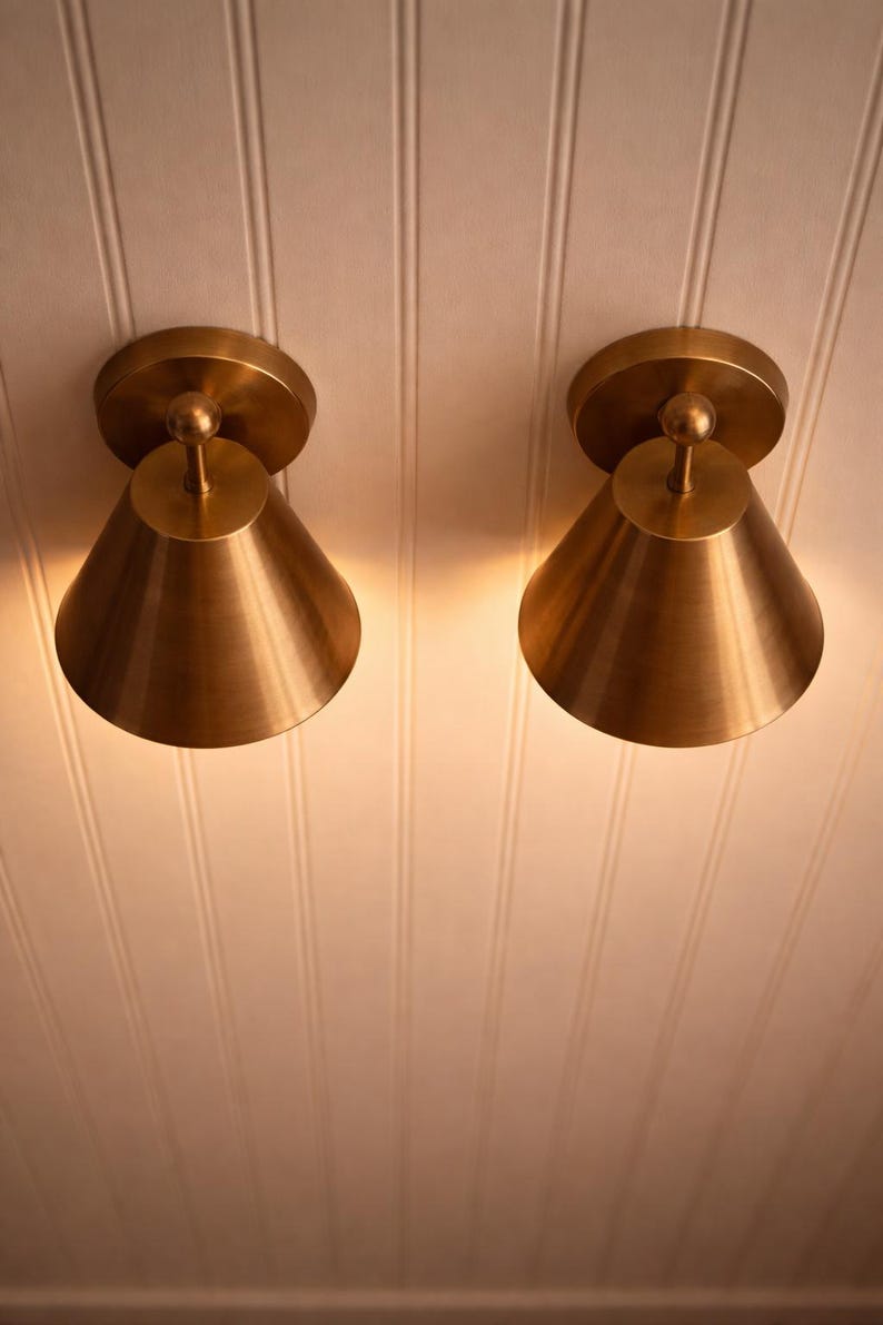 May include: Two brass-colored cone-shaped wall sconces mounted on a white paneled ceiling. Each sconce has a circular base and a small, spherical detail at the top. The sconces are illuminated, casting a warm glow.