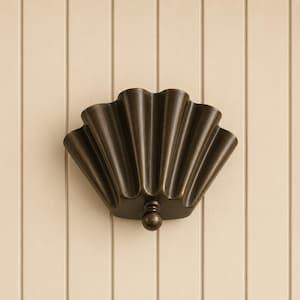 May include: A dark bronze-toned wall sconce featuring a fan-shaped shell design with vertical fluted ridges. The fixture has a rounded knob at the base and a smooth, metallic finish, providing a structured and symmetrical appearance for wall lighting.