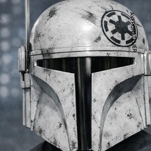 May include: A distressed white Mandalorian helmet with black detailing and a black visor. The helmet displays a circular emblem and the text "AVERONATIER". A black antenna projects from the top. The helmet has a worn, battle-damaged look.
