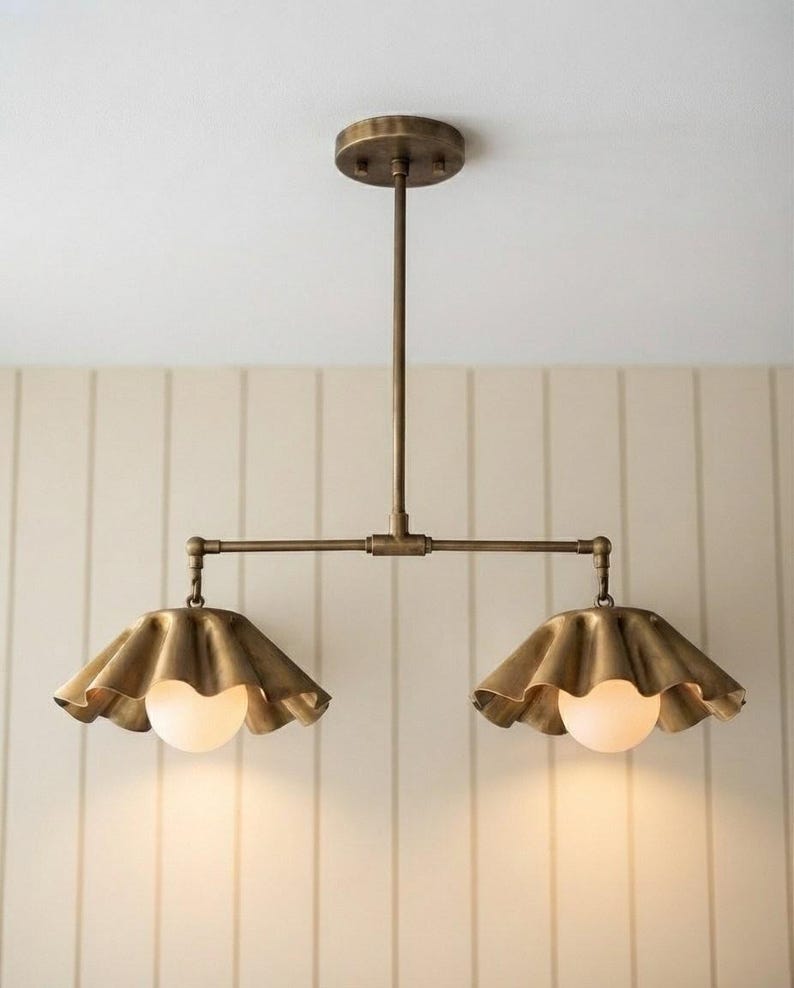 May include: A dual-light pendant fixture featuring a brass-toned metal finish with a central vertical rod and a horizontal bar. Two ruffled, scalloped-edge metal shades hang from the ends, each housing a round, frosted white glass light bulb.